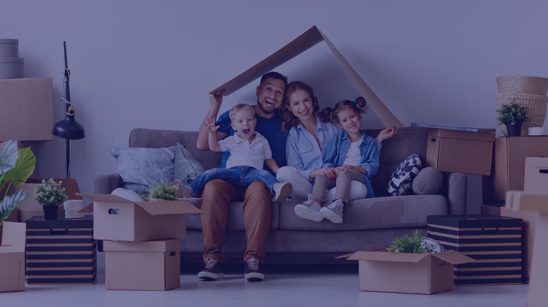A family of four sits on a couch surrounded by moving boxes, holding a cardboard piece shaped like a roof above them.