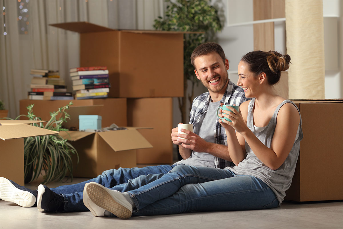 A man and a woman sit on the floor, drinking from mugs They are surrounded by moving boxes and a potted plant