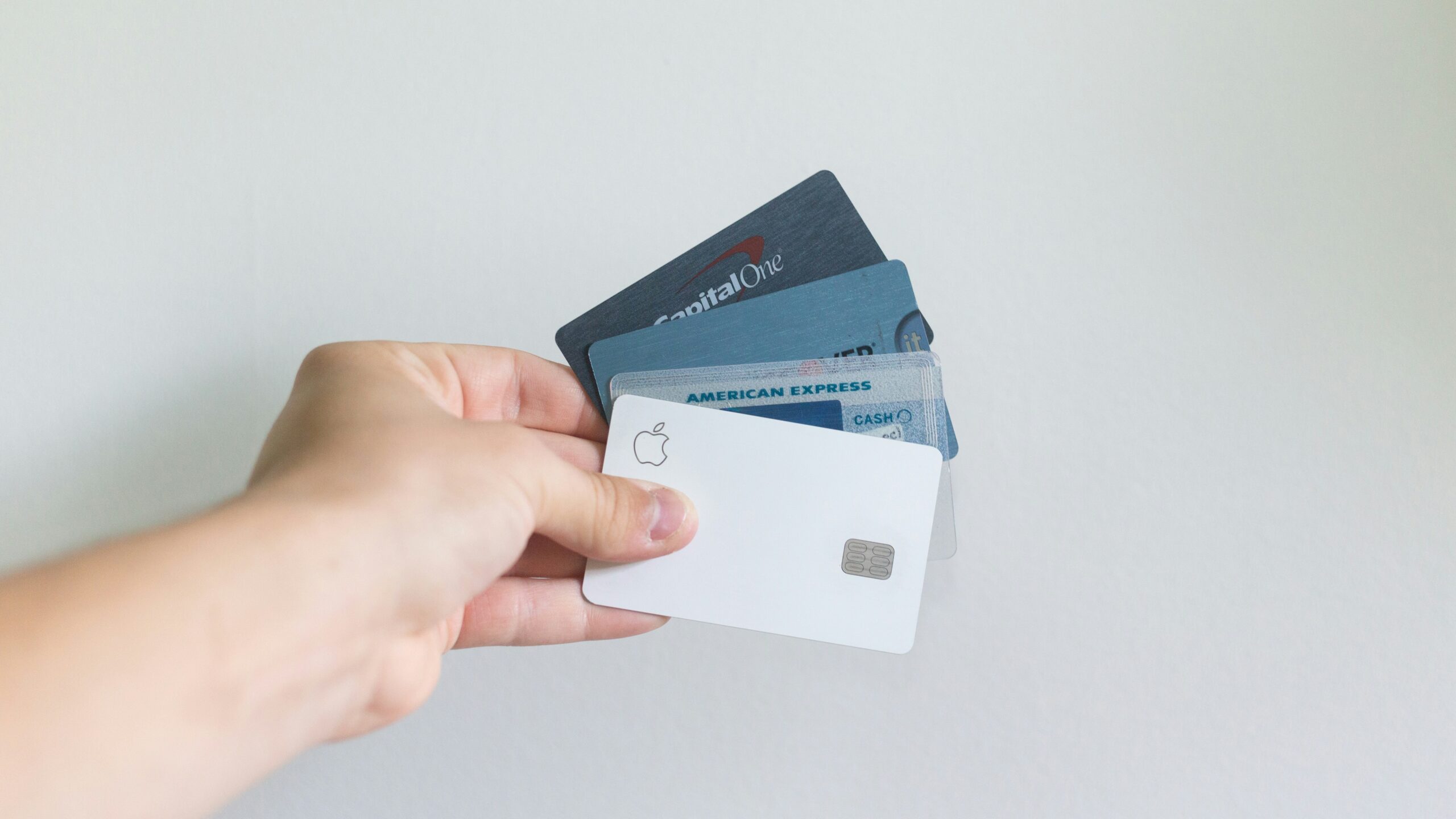 Hand holding four credit cards, including an Apple Card and an American Express, against a plain background